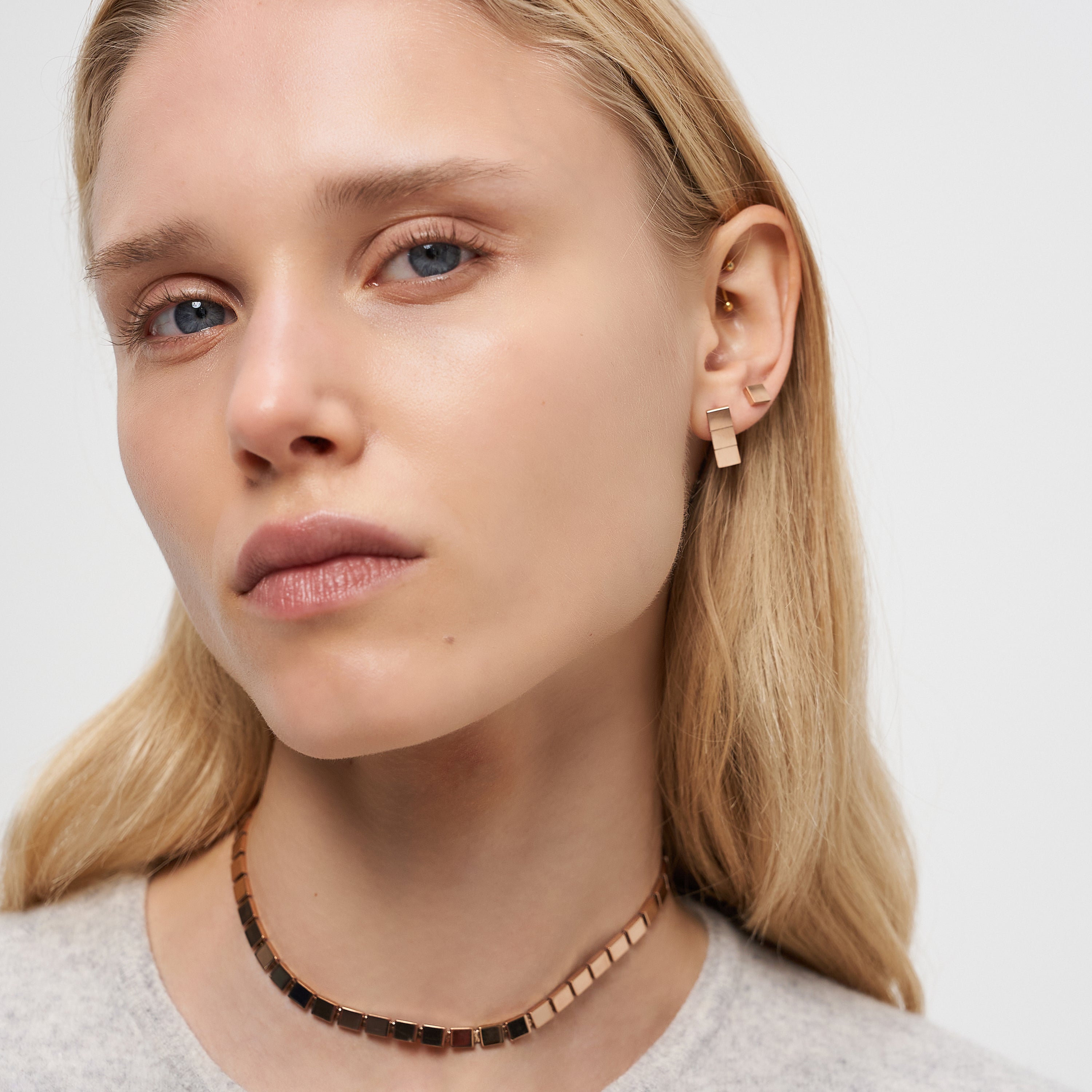 Frau mit glattem blondem Haar trägt einen goldenen Liebeskind Berlin Ohrring - Plates und eine Halskette, schaut mit neutralem Blick in die Kamera und setzt auf minimalistisches Design.