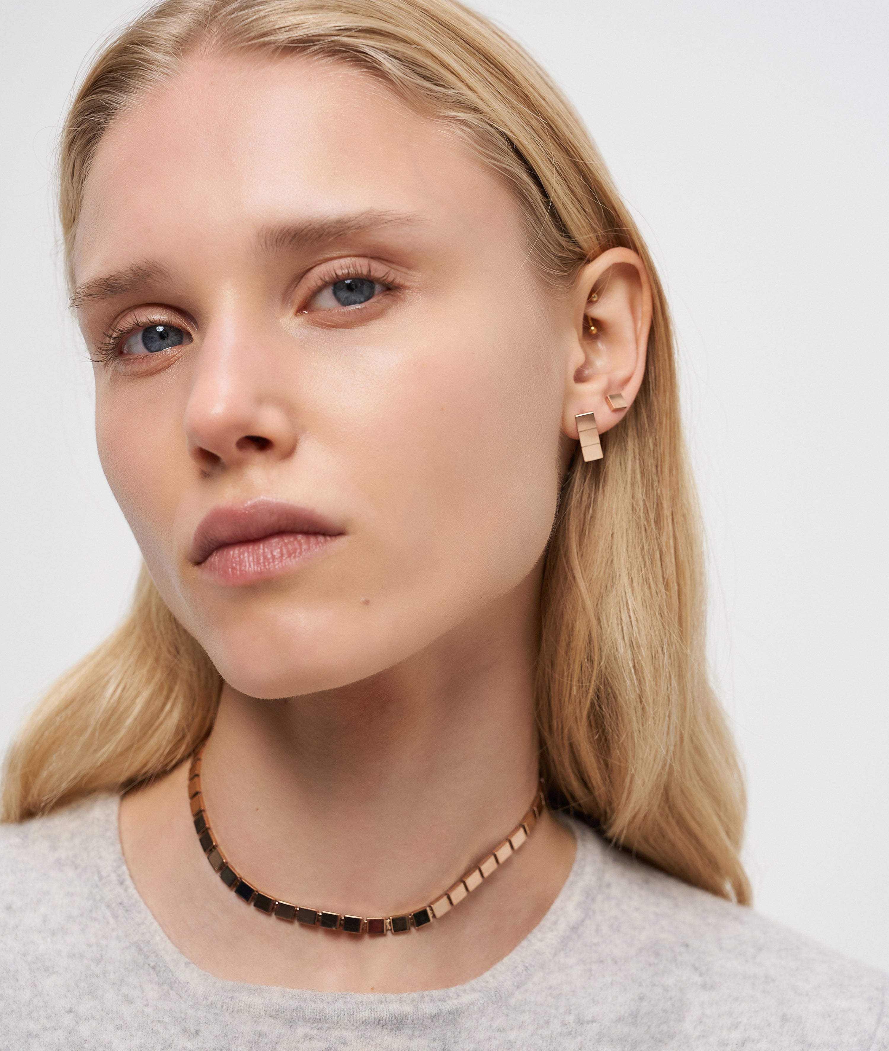 Frau mit glattem blondem Haar trägt einen goldenen Liebeskind Berlin Ohrring - Plates und eine Halskette, schaut mit neutralem Blick in die Kamera und setzt auf minimalistisches Design.