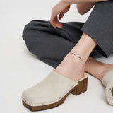 Eine Person in grauer Hose und beigen Clogs mit Absatz sitzt auf einem weißen Boden und trägt die elegante Liebeskind Berlin Fußkette - The Sleek an ihrem Knöchel.