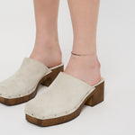 Eine Person modelliert beigefarbene Wildleder-Plateau-Clogs mit Holzabsatz und silbernen Nieten, gepaart mit der Liebeskind Berlin Fußkette - The Sleek von Liebeskind Berlin, vor einem weißen Hintergrund.