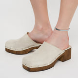 Person trägt beige Wildlederclogs mit Holzabsatz und silbernen Nieten, gepaart mit der Liebeskind Berlin Fußkette - The Sleek an einem Knöchel.