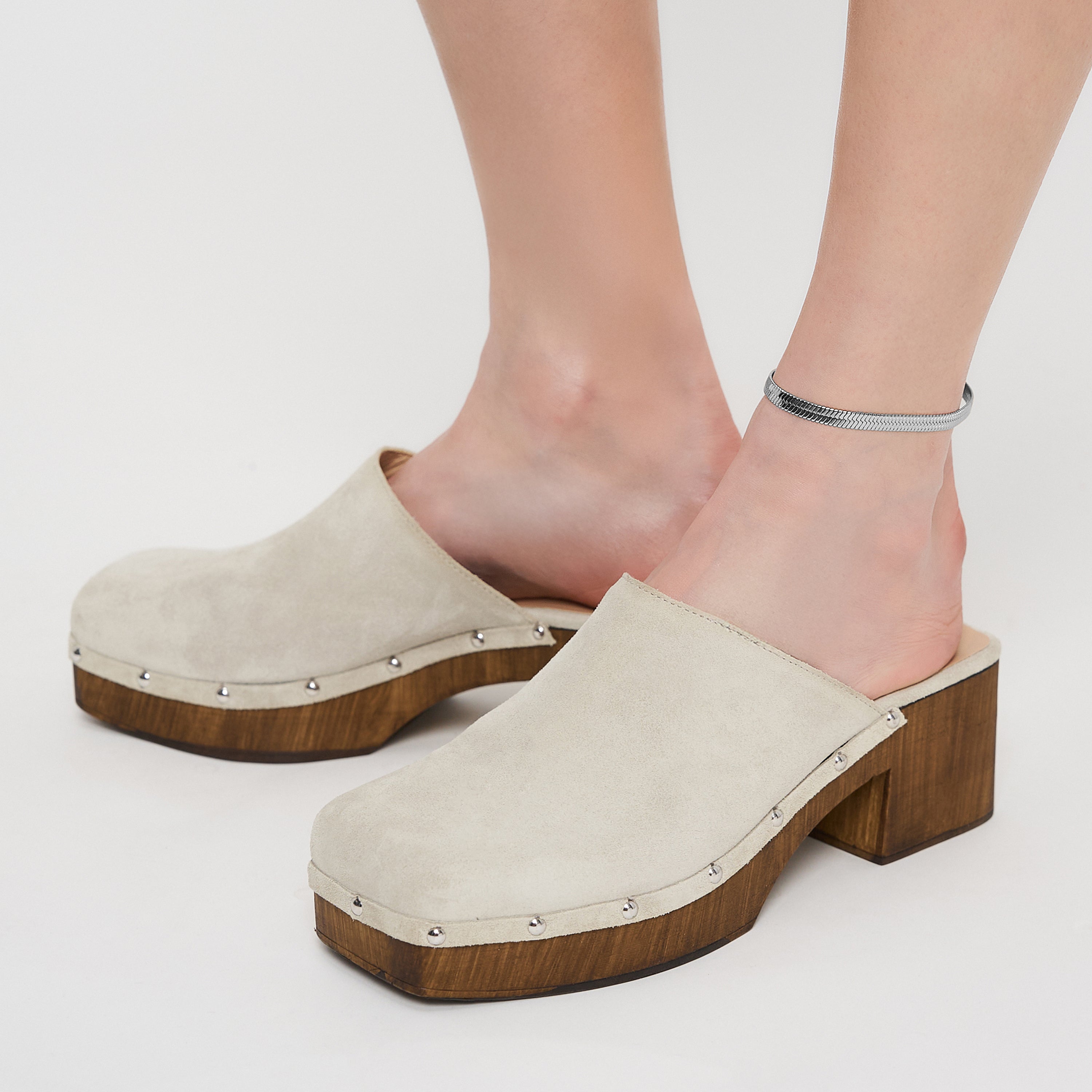Person trägt beige Wildlederclogs mit Holzabsatz und silbernen Nieten, gepaart mit der Liebeskind Berlin Fußkette - The Sleek an einem Knöchel.
