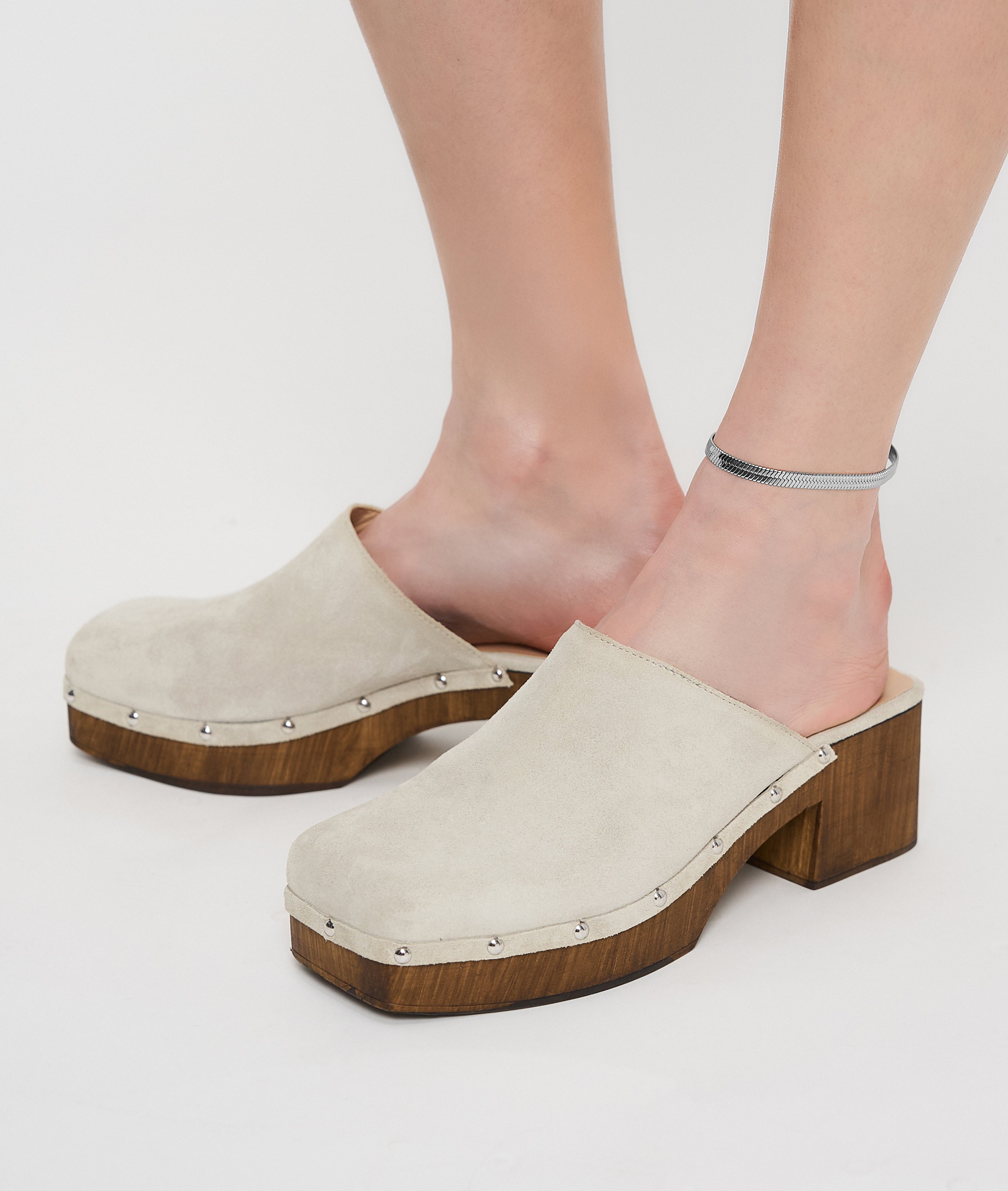 Person trägt beige Wildlederclogs mit Holzabsatz und silbernen Nieten, gepaart mit der Liebeskind Berlin Fußkette - The Sleek an einem Knöchel.