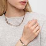 Eine Frau in einem hellgrauen Pullover, die Hand am Kragen, präsentiert die Liebeskind Berlin Halskette - The Sleek, gepaart mit einem passenden Silberarmband.