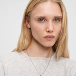 Eine Frau mit langen blonden Haaren trägt einen leichten Pullover und eine Liebeskind Berlin Halskette - Broken Heart mit einem zarten Silberanhänger.