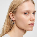 Frau mit glattem blondem Haar trägt zwei silberne Liebeskind Berlin Ohrring - Organic Shapes Ohrringe, einer mit einem kleinen Herzanhänger, der zur Seite schaut.