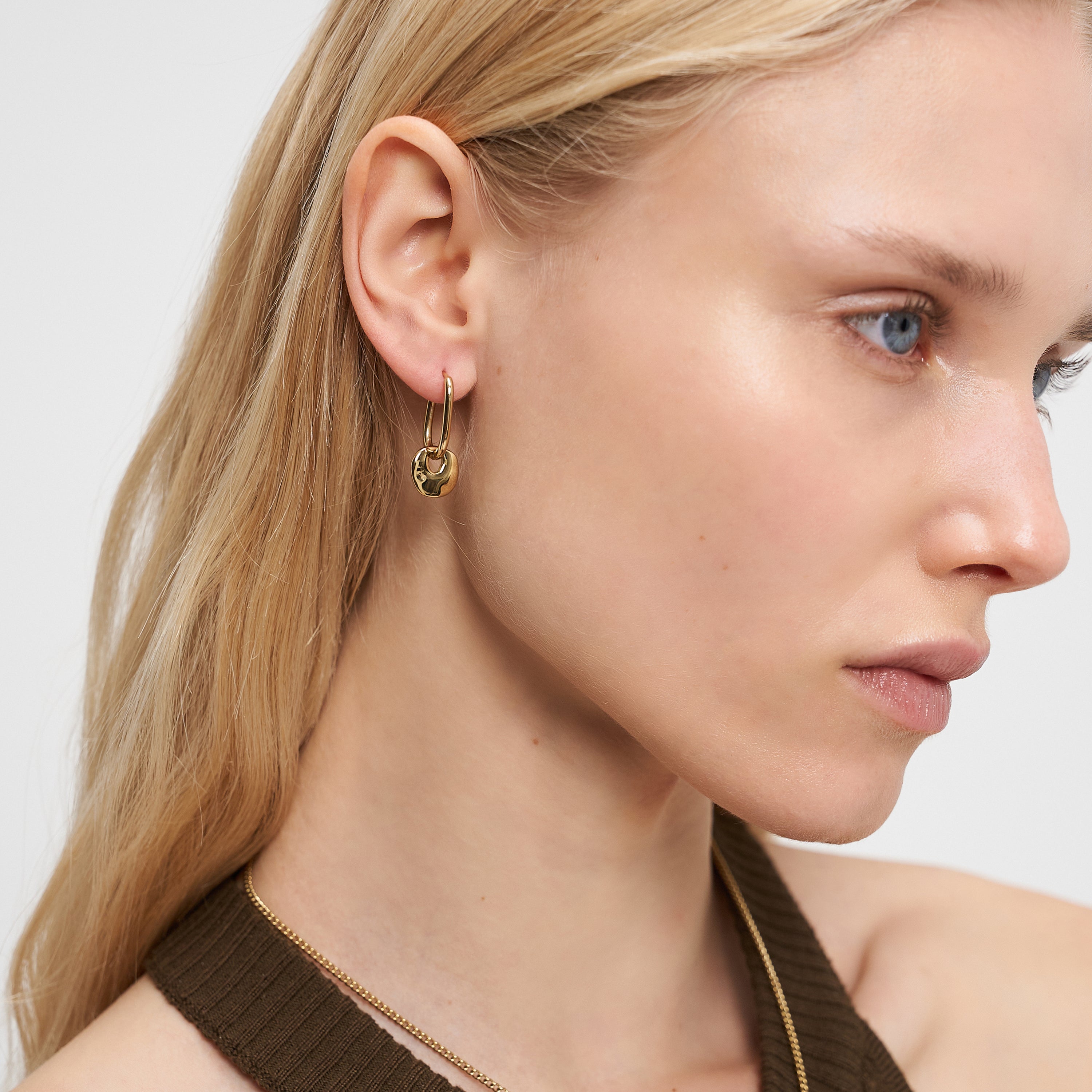 Frau mit langen blonden Haaren trägt Liebeskind Berlin Ohrring - Organic Shapes, goldene Creolen von Liebeskind Berlin, gepaart mit einem braunen Neckholder-Top, von rechts.