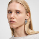 Eine Frau mit glattem blondem Haar trägt den Liebeskind Berlin Ohrring - Organic Shapes und einen hellgrauen Pullover und schaut in die Kamera.