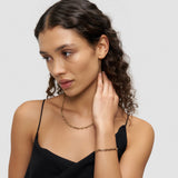 Frau mit lockigem Haar trägt ein schwarzes Oberteil, eine goldene Halskette und das Liebeskind Berlin LIEBESKIND BERLIN Armband - Urban Chic, mit Blick zur Seite.