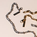 Das LIEBESKIND BERLIN Armband - Urban Chic von Liebeskind Berlin verfügt über verstellbare Silber- und Goldketten mit STAINLESS STEEL-Anhängern auf einer cremefarbenen Oberfläche und verbindet urbanes Design mit persönlicher Anpassung.