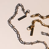 Das LIEBESKIND BERLIN Armband - Urban Chic von Liebeskind Berlin verfügt über verstellbare Silber- und Goldketten mit STAINLESS STEEL-Anhängern auf einer cremefarbenen Oberfläche und verbindet urbanes Design mit persönlicher Anpassung.