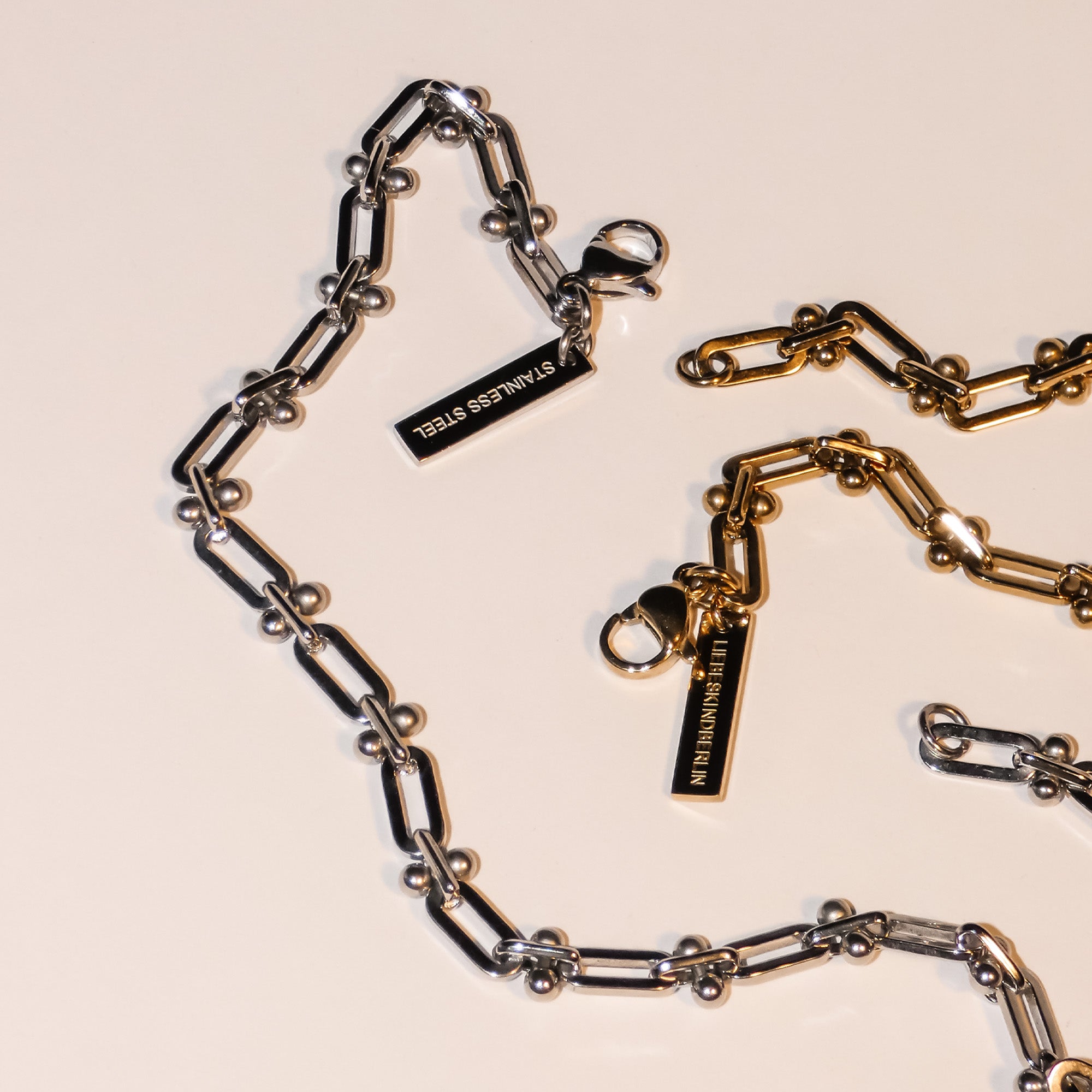 Das LIEBESKIND BERLIN Armband - Urban Chic von Liebeskind Berlin verfügt über verstellbare Silber- und Goldketten mit STAINLESS STEEL-Anhängern auf einer cremefarbenen Oberfläche und verbindet urbanes Design mit persönlicher Anpassung.