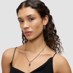 Eine Frau mit lockigem Haar trägt ein schwarzes Oberteil, Liebeskind Berlin Halskette - Metal Heart, und passende goldene Ohrringe.