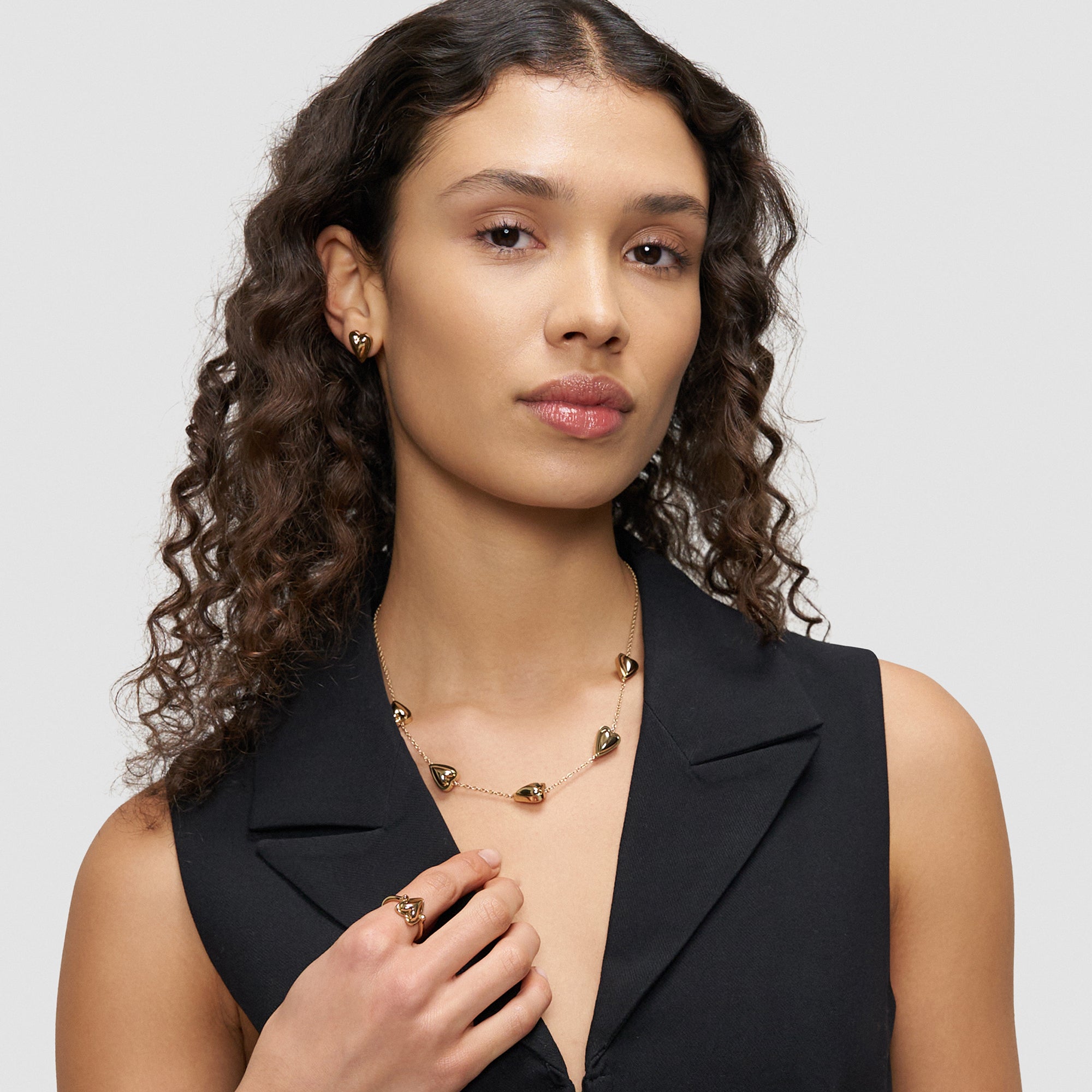 Frau mit langen lockigen Haaren trägt ein schwarzes ärmelloses Oberteil und eine goldene Liebeskind Berlin Halskette - Metal Heart und posiert vor einem schlichten Hintergrund.
