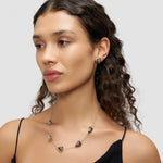 Eine Frau mit lockigem Haar trägt eine Herz-Design Halskette in Herzform und den dazu passenden LIEBESKIND BERLIN Ohrstecker - Metal Heart von Liebeskind Berlin, gepaart mit einem schwarzen Oberteil vor grauem Hintergrund.