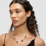 Eine Frau mit lockigem Haar trägt eine Herz-Design Halskette in Herzform und den dazu passenden LIEBESKIND BERLIN Ohrstecker - Metal Heart von Liebeskind Berlin, gepaart mit einem schwarzen Oberteil vor grauem Hintergrund.