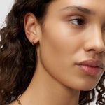 Eine Frau mit gewelltem Haar, die zur Seite schaut, trägt einen kleinen Liebeskind Berlin Creole - Metal Heart goldenen Creolenohrring und eine Halskette mit einem Herzanhänger.