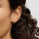 Nahaufnahme eines Frauenohrs mit lockigem Haar, das zwei Liebeskind Berlin Creole - Metal Heart Ohrringe und eine silberne Ohrmanschette trägt, die urbanes Design präsentieren.