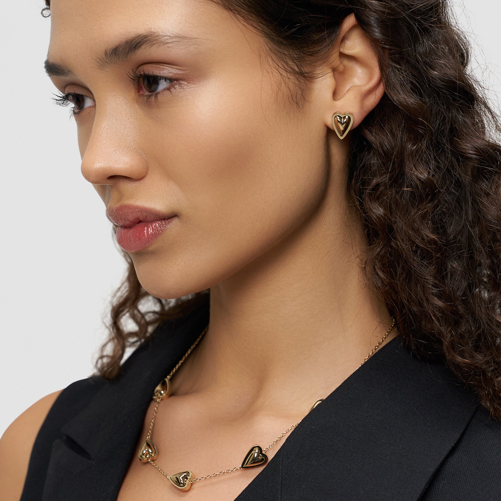 Eine Frau mit lockigem Haar trägt den LIEBESKIND BERLIN Ohrstecker - Metal Heart von Liebeskind Berlin und eine dazu passende Herz-Halskette. Sie präsentiert eleganten Schmuck im klassischen Herz-Design, gekleidet in Schwarz.