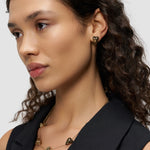 Frau mit lockigem Haar trägt LIEBESKIND BERLIN Ohrstecker - Metal Heart von Liebeskind Berlin und goldenen herzförmigen Schmuck, gestylt mit einem schwarzen ärmellosen Top.