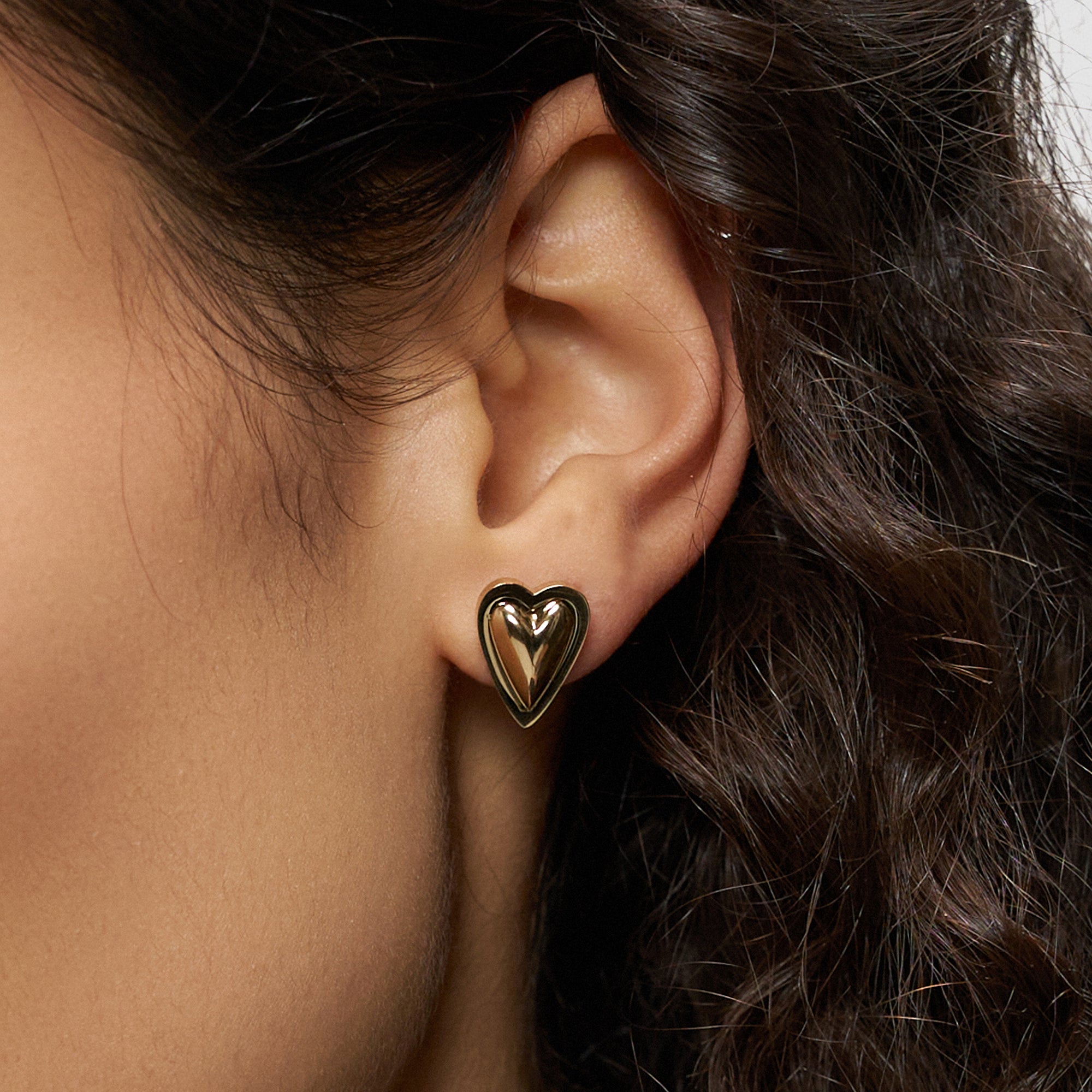 Nahaufnahme eines Frauenohrs, das den Liebeskind Berlin Ohrstecker - Metal Heart in glänzendem Gold trägt. Das elegante Herzdesign wird durch ihr lockiges braunes Haar wunderschön hervorgehoben und verleiht ihr einen romantischen Touch.