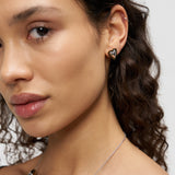 Frau mit lockigem Haar, die einen kleinen Liebeskind Berlin Ohrstecker - Metal Heart und eine silberne Halskette trägt, schaut in die Kamera.