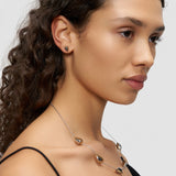 Eine Frau mit lockigem Haar blickt zur Seite und trägt eine tropfenförmige Halskette und passende Liebeskind Berlin Ohrstecker - Metal Heart Ohrringe - ein elegantes Schmuckensemble.