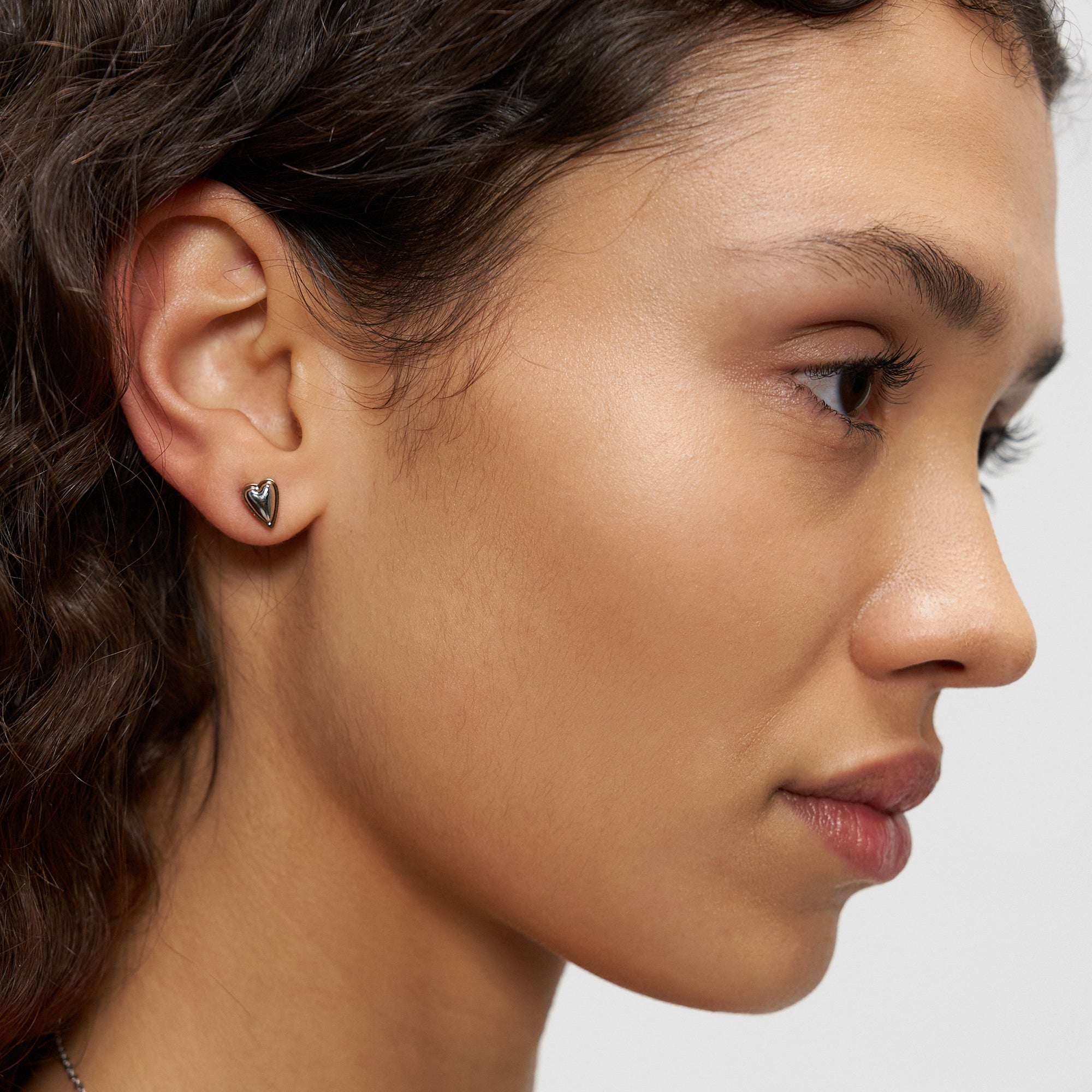 Eine Frau mit lockigem Haar trägt einen kleinen Liebeskind Berlin Ohrstecker - Metal Heart in ihrem linken Ohr, dargestellt in einer Nahaufnahme im Seitenprofil.