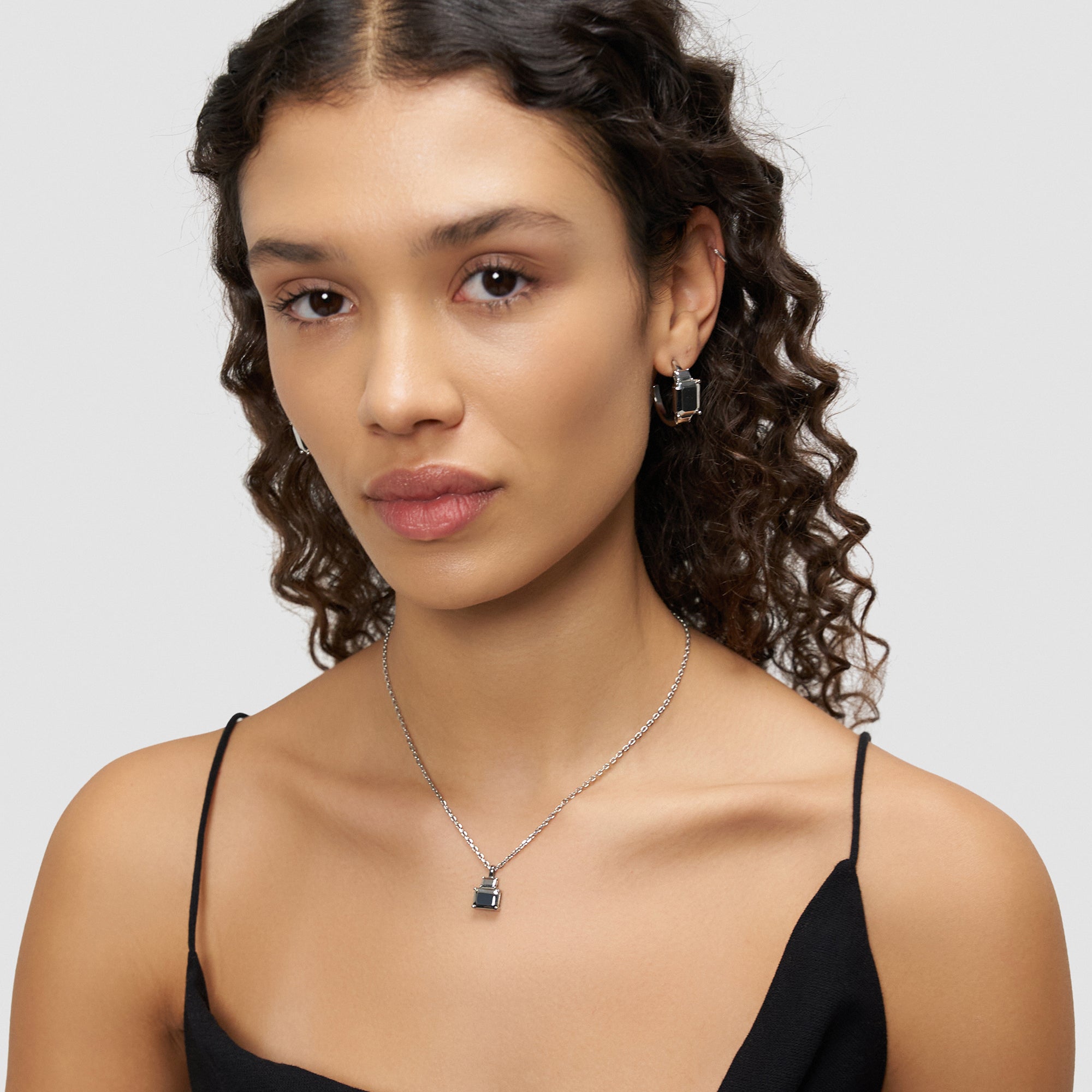 Frau mit lockigem Haar, die ein schwarzes Oberteil und die Liebeskind Berlin Halskette - Steel Diamond sowie passende Ohrringe trägt und in die Kamera schaut.