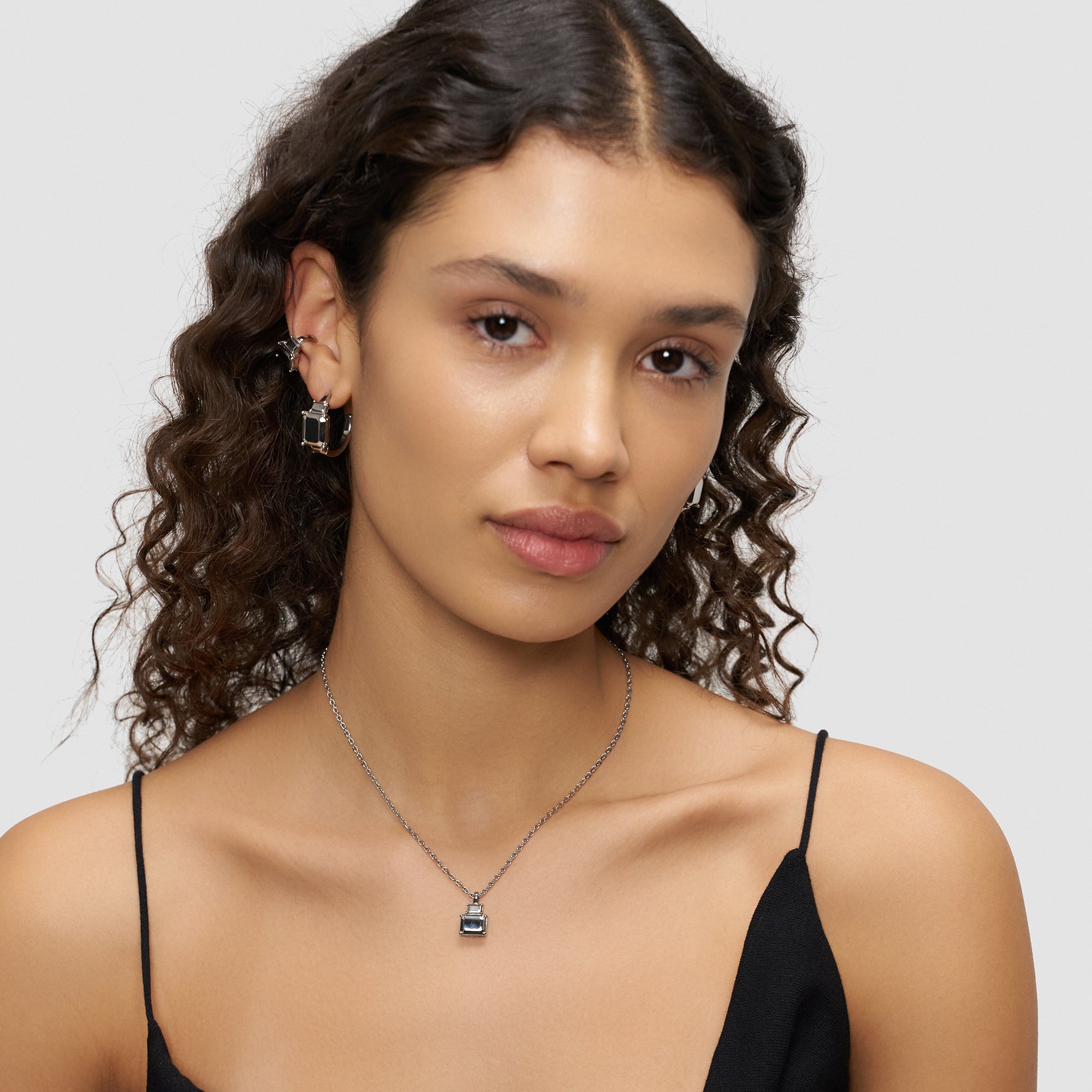 Frau mit lockigem Haar trägt ein schwarzes Oberteil, Ohrringe und die Liebeskind Berlin Halskette - Steel Diamond, vor einem hellgrauen Hintergrund.