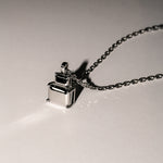 Die LIEBESKIND BERLIN Halskette - Steel Diamond von Liebeskind Berlin mit ihrem geometrischen, minimalistischen Anhänger wirft einen Schatten auf eine schlichte Oberfläche.