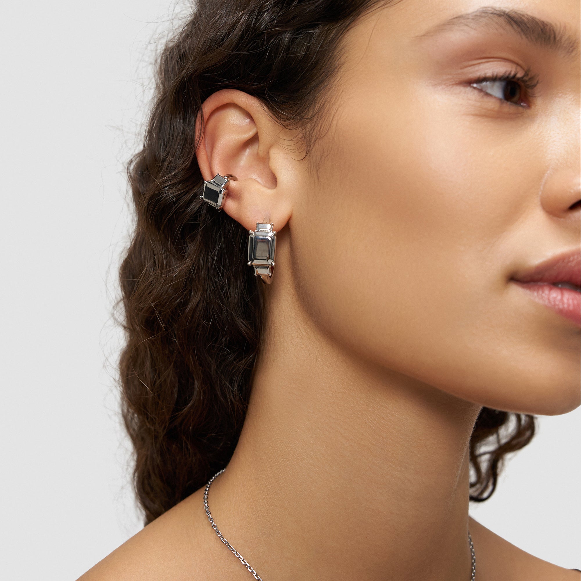 Eine Frau mit lockigem Haar trägt zwei silberne Ohrringe und eine Halskette, im Profil gezeigt, gestylt mit der LIEBESKIND BERLIN Earcuff - Steel Diamond von Liebeskind Berlin.