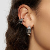 Nahaufnahme eines Frauenohrs, das zwei silberne Ohrringe mit rechteckigen schwarzen Edelsteinen in Diamantoptik und einen LIEBESKIND BERLIN Earcuff - Steel Diamond von Liebeskind Berlin trägt.