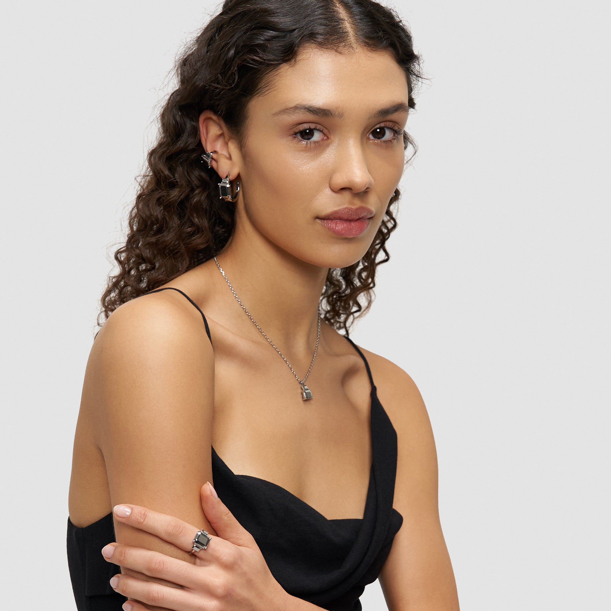 Frau mit lockigem Haar, die ein schwarzes Kleid, eine silberne Halskette, Ohrringe und den Liebeskind Berlin Ring - Steel Diamond in 54 trägt, posiert vor einem schlichten Hintergrund.
