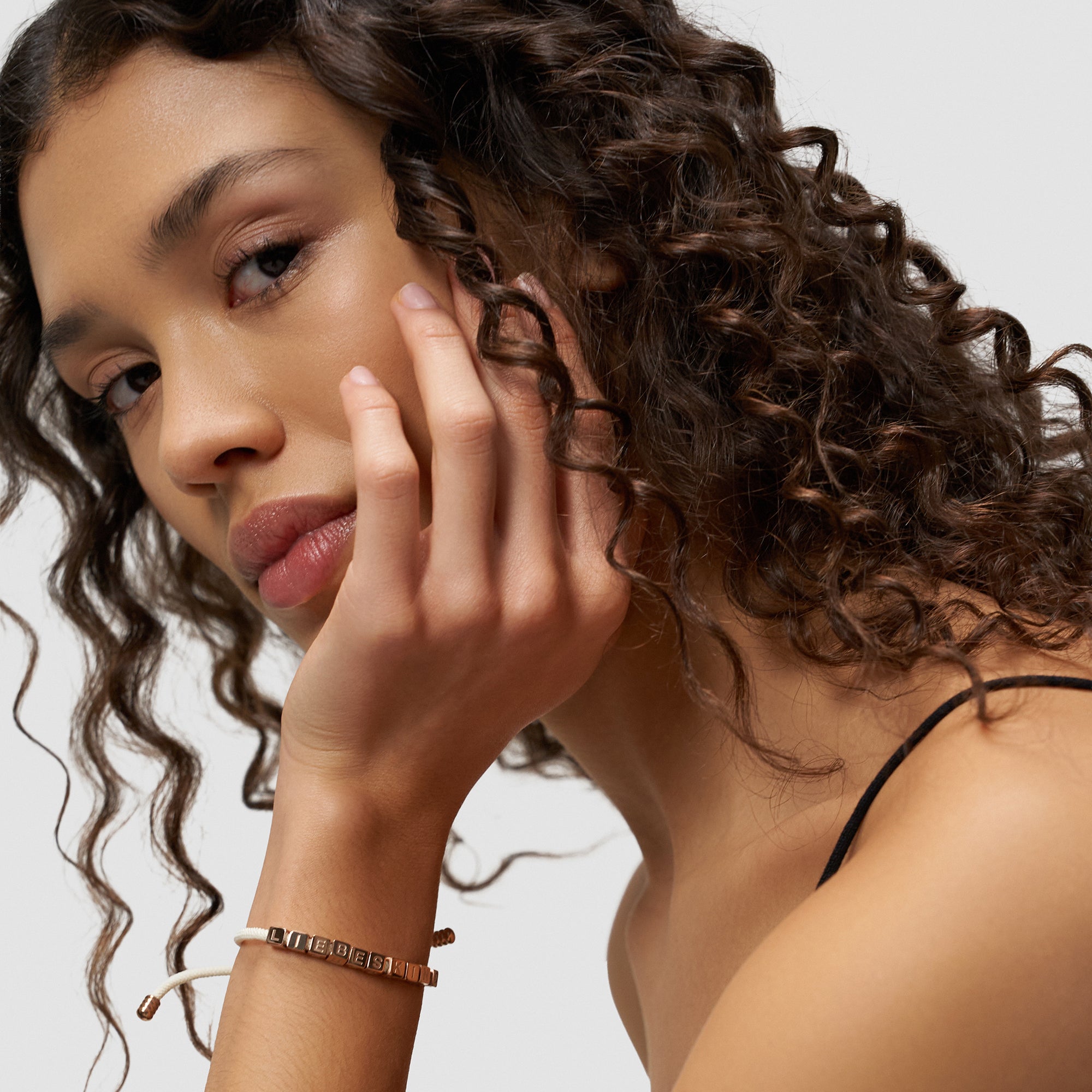 Eine Frau mit lockigem Haar stützt ihr Kinn auf die Hand. Sie trägt ein schwarzes Trägertop und das Liebeskind Berlin Armband - Brick als gewagtes Statement Piece.