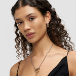 Frau mit lockigem Haar trägt ein schwarzes Oberteil, goldene Ohrringe und die Liebeskind Berlin Halskette - Urban Chic-perfekt für Liebhaber von zeitlosem Design.
