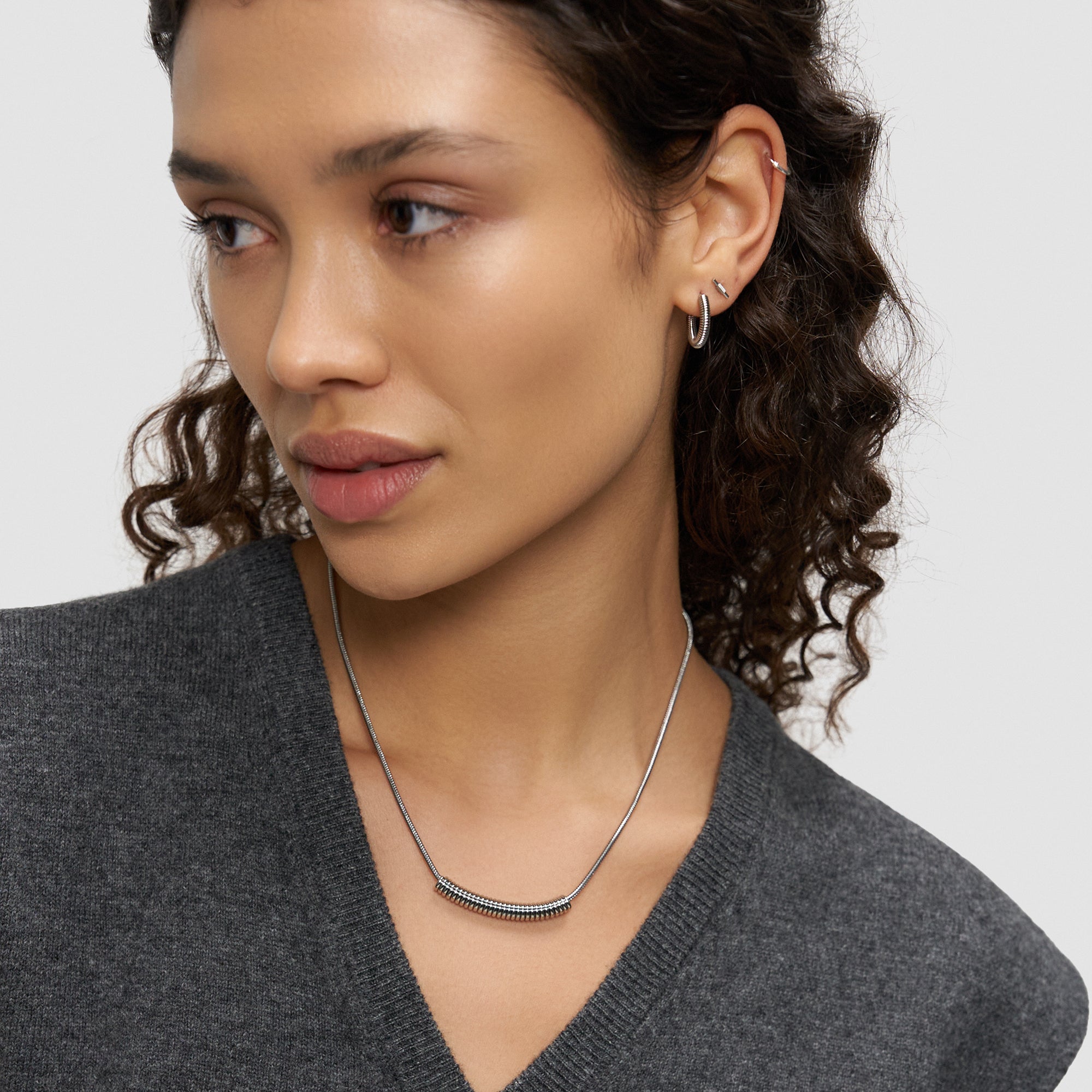 Eine Frau mit lockigem Haar, die ein graues Oberteil, silberne Ohrringe und die Liebeskind Berlin Halskette - Twist trägt, schaut zur Seite.