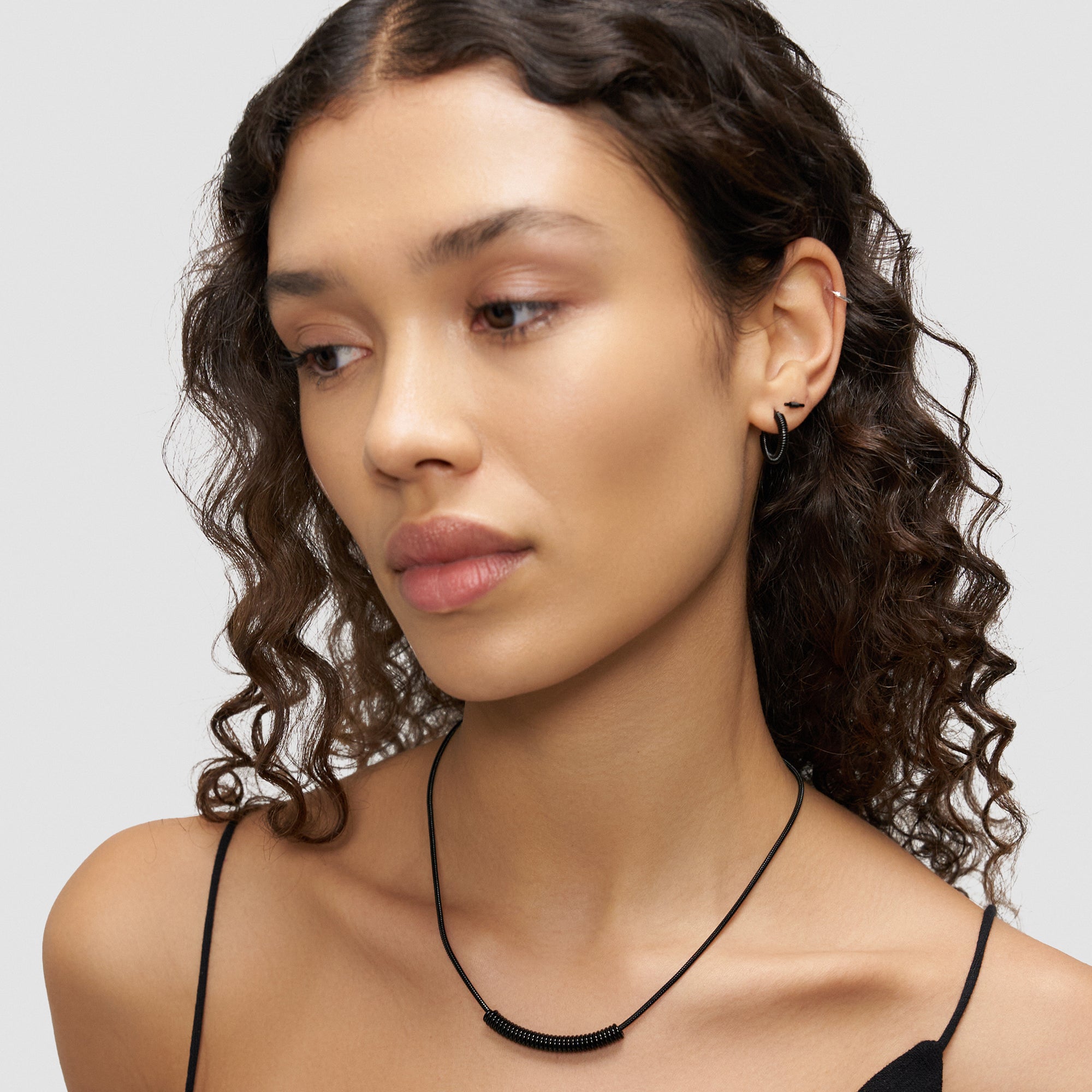 Frau mit lockigem Haar, die die LIEBESKIND BERLIN Halskette - Twist von Liebeskind Berlin und Ohrringe trägt und in einem schwarzen Spaghettiträgertop nach unten schaut.