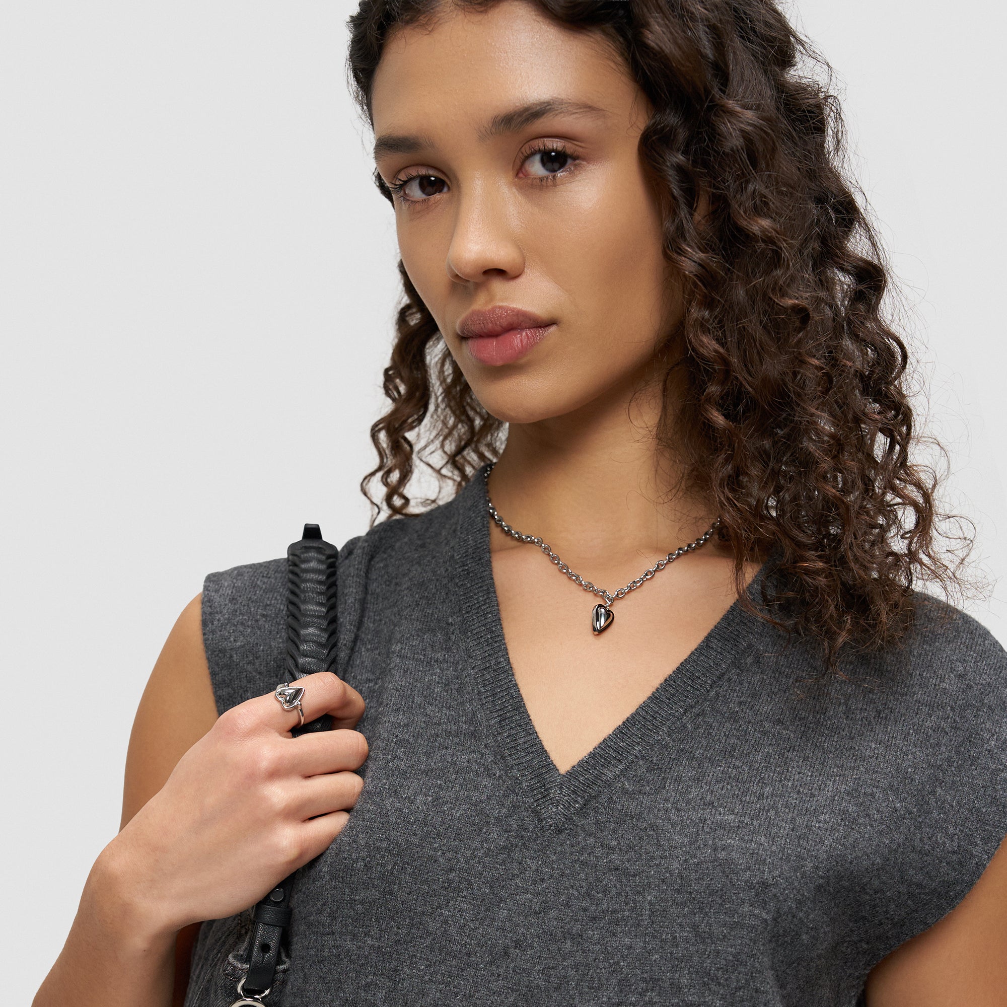 Eine Frau mit lockigem Haar in einem grauen ärmellosen Pullover trägt eine Halskette und den Liebeskind Berlin Ring - Metal Heart in 54, der ein schwarzes Armband hält - ein stilvoller Look, der sich perfekt als Geschenk eignet.