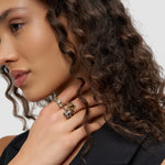 Eine Frau mit lockigem Haar und schwarzem Oberteil präsentiert den Liebeskind Berlin Glasherz-Ring in Größe 52 - ein echtes Statement - in Kombination mit Silberschmuck an ihrer Hand.