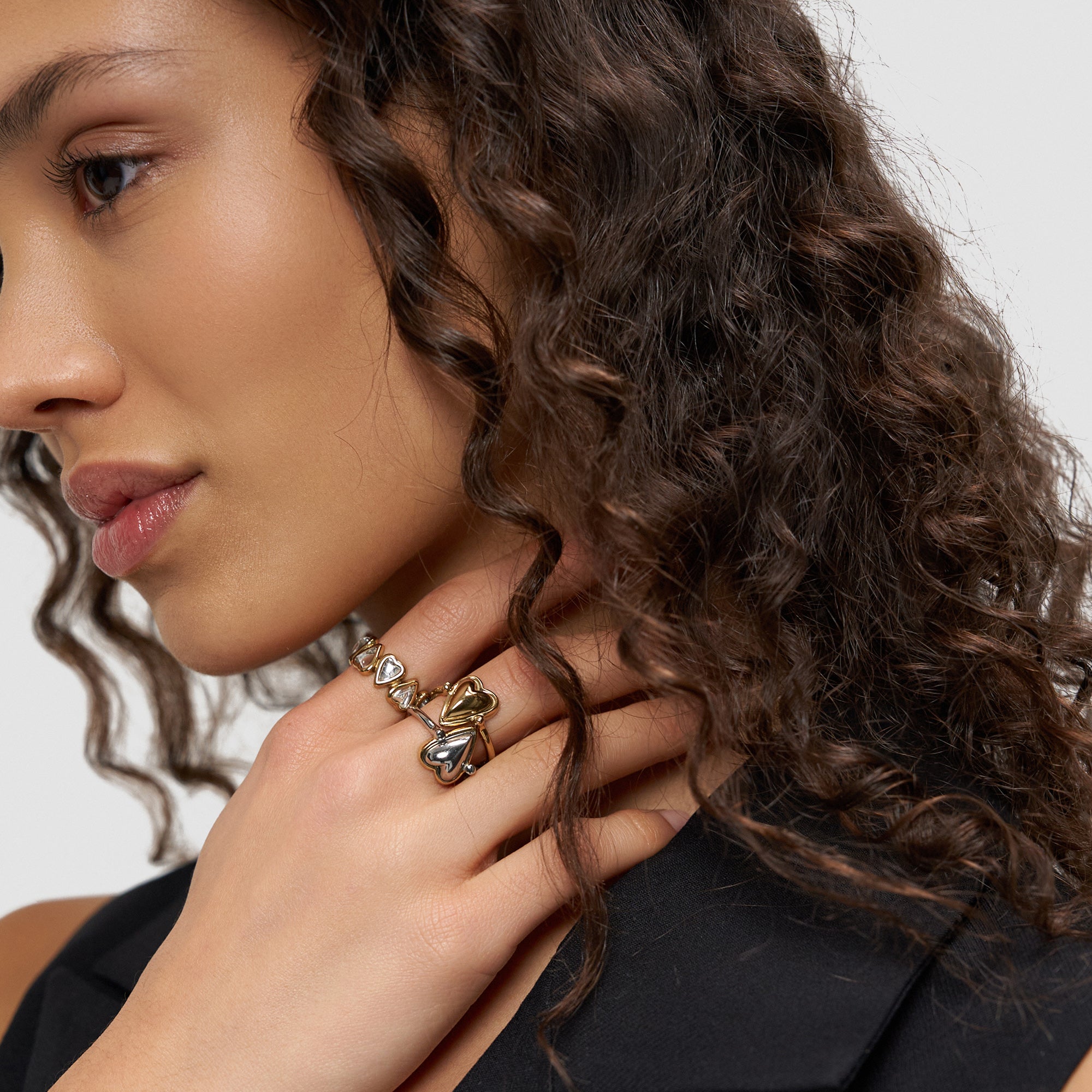 Eine Frau mit lockigem Haar und schwarzem Oberteil präsentiert den Liebeskind Berlin Glasherz-Ring in Größe 52 - ein echtes Statement - in Kombination mit Silberschmuck an ihrer Hand.