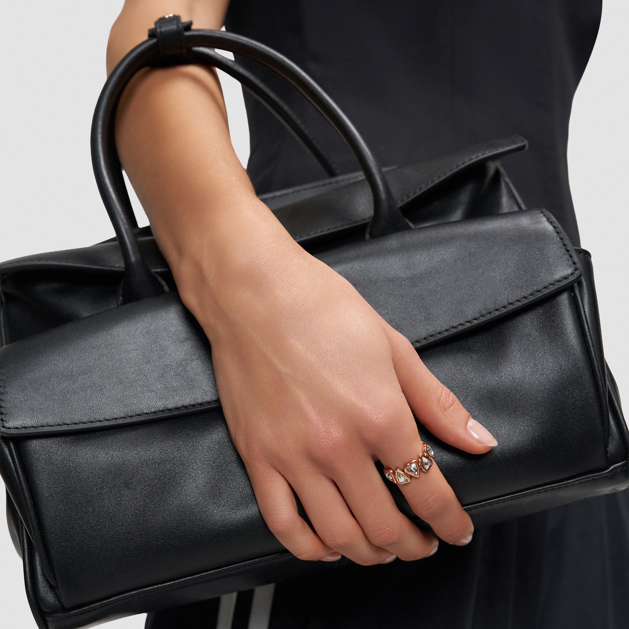 Eine mit dem Liebeskind Berlin Ring - Glass Heart in 54 geschmückte Hand hebt eine schwarze Lederhandtasche von einem dunklen Outfit ab und lässt beide Liebeskind Berlin Accessoires hervorstechen.