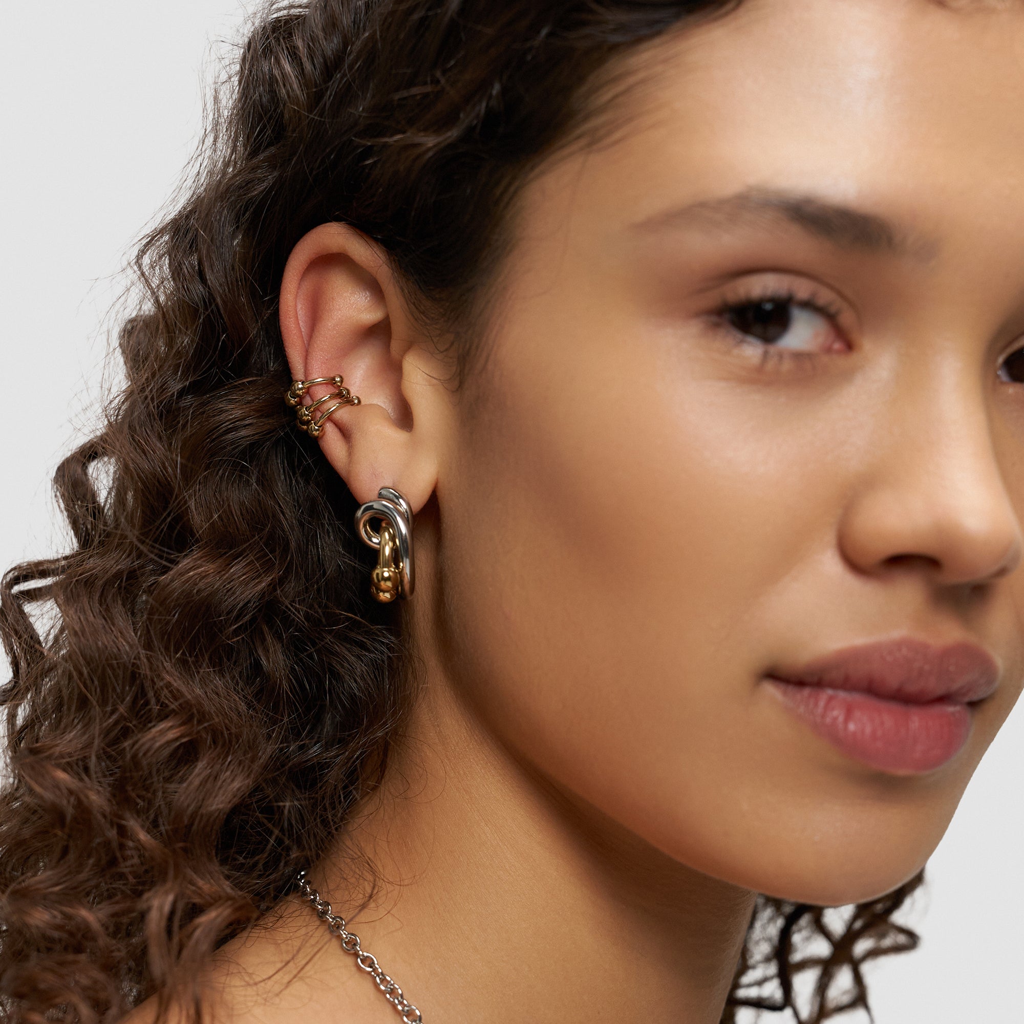 Eine Frau mit lockigem Haar trägt mehrere Ohrringe - darunter goldene und silberne Creolen sowie das LIEBESKIND BERLIN Earcuff Set - Urban Chic by Liebeskind Berlin - während sie in die Kamera schaut.