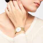 Eine Person, die die Liebeskind Berlin Set - Golden Green Uhr mit Netzband trägt, berührt sanft ihren Hals, bekleidet mit einem cremefarbenen Oberteil.