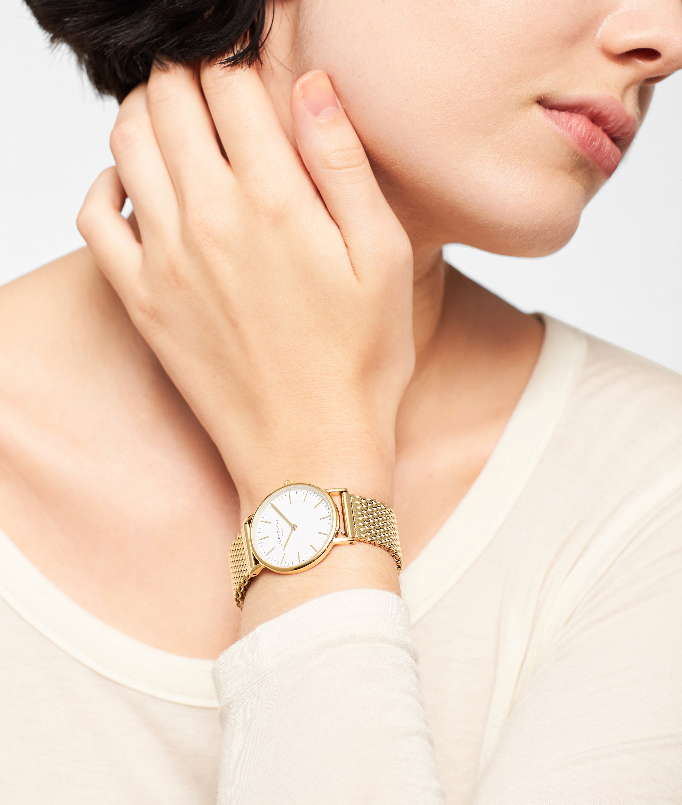 Eine Person, die die Liebeskind Berlin Set - Golden Green Uhr mit Netzband trägt, berührt sanft ihren Hals, bekleidet mit einem cremefarbenen Oberteil.