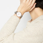 Person, die einen beigen Pullover und die LIEBESKIND BERLIN Armbanduhr - The Vintage Must-Have von Liebeskind Berlin mit minimalistischem Design und hellgrauem Armband trägt, wobei die Hand den Kopf berührt.