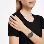 Eine Frau in einem schwarzen T-Shirt trägt das Liebeskind Berlin Set - Lovely Silver Basic mit einer eleganten Uhr mit schwarzem Zifferblatt und einem eleganten Herzarmband für einen mühelos schicken Look.