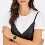 Eine Frau in weißem T-Shirt und schwarzem Kleid trägt die LIEBESKIND BERLIN Armbanduhr - The Colored Scandinavian mit grünem Ziffernblatt und Edelstahl-Mesh-Armband, die das moderne skandinavische Design von Liebeskind Berlin unterstreicht.