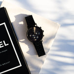 Eine LIEBESKIND BERLIN Armbanduhr - Modern Chronograph in Schwarz ruht auf cremefarbenem Stoff neben einem monochromen Buch, dessen sanfte Schatten ihre moderne Eleganz unterstreichen - ideal für alle, die eine stilvolle Damenuhr von Liebeskind Berlin suchen.