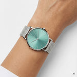 Ki generiertes Bild einer LIEBESKIND BERLIN Damenuhr mit silberfarbenem Edelstahl-Mesharmband, silberfarbenem Gehäuse und türkisfarbenem Sunray-Zifferblatt, getragen am Handgelenk.